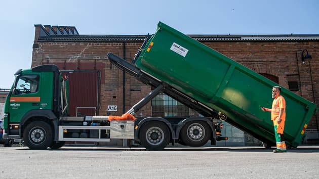 En man i varselkläder lastar av en container från en lastbil med en fjärrkontroll. Bakom syns en tegelbyggnad och blå himmel.