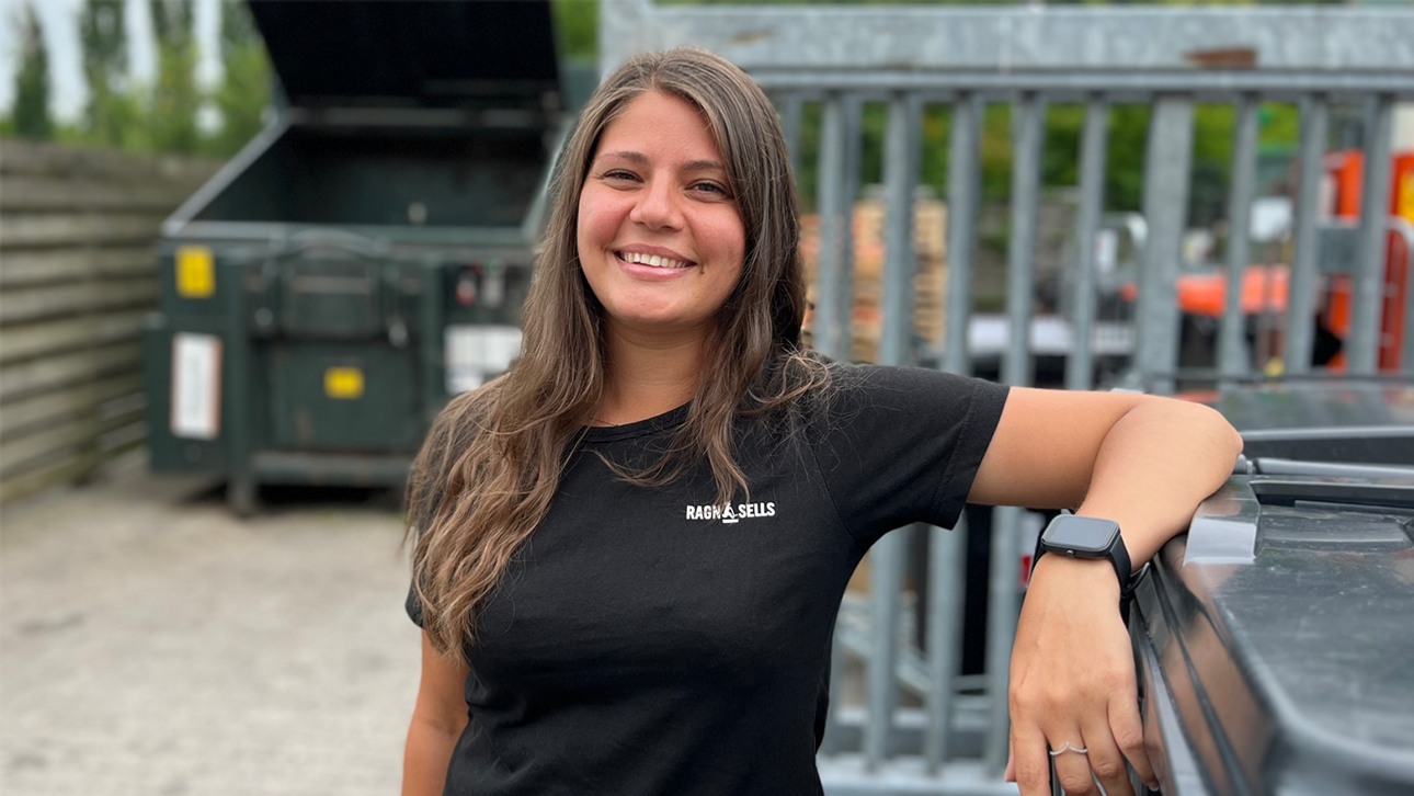 En tjej med svart t-shirt där det står Ragn-Sells som luta sig mot ett avfallskärl