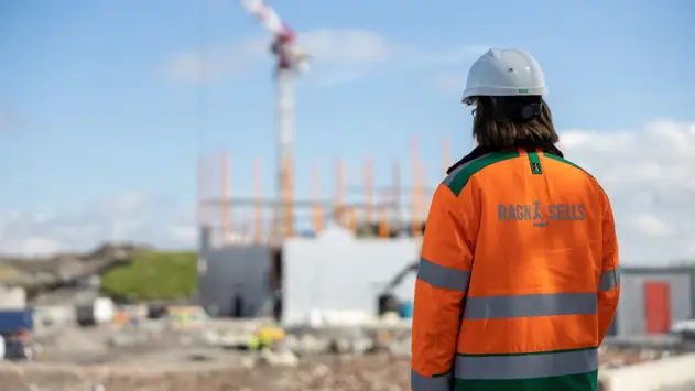 en man från Ragn-Sells står med ryggen mot i orange jacka och tittar på ett stort bygge