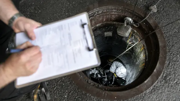 En hand med ett kontrollpapper ovanför ett hål (oljeavskiljare) där en kontrollant är nere och inspekterar