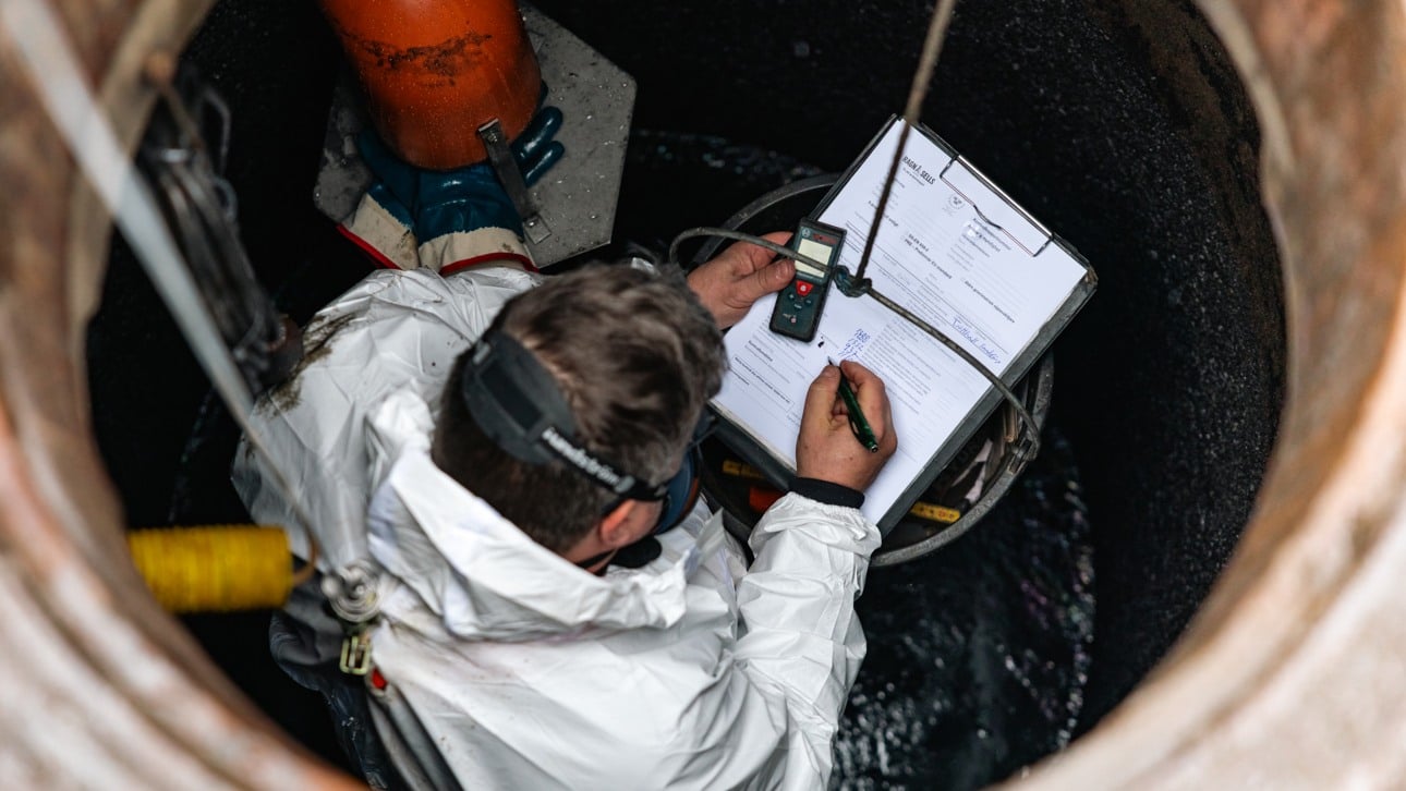 Man fr&aring;n Ragn-Sells i vit skyddsdr&auml;kt och munskydd nere i en Oljeavskiljare och fyller i protokoll