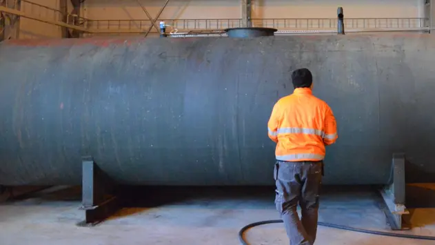 En man bakifrån med orange jacka som står framför en stor grå cistern inomhus
