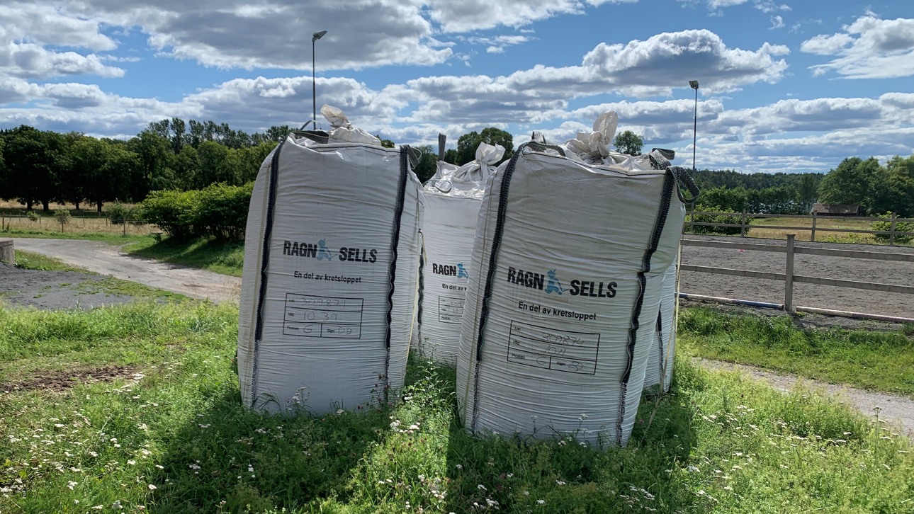 Tre fulla byggsäckar på en gräsplätt med en hage bakom och blå himmel med lite moln